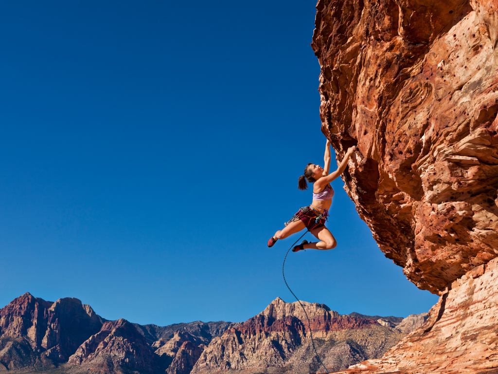 Woman rock climbing on red mountains with just a tether around her waste and hanging on rock fron by hands feet outstretched